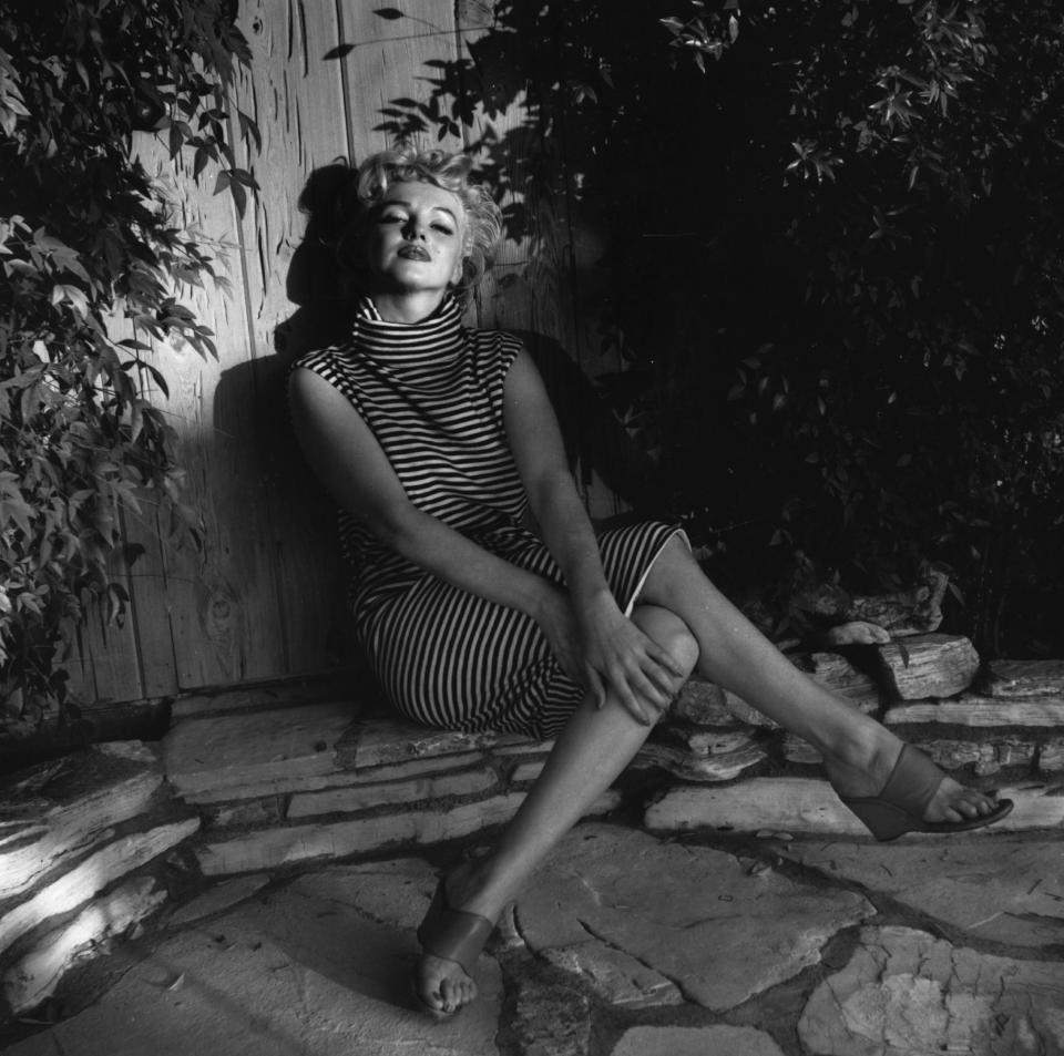 Marilyn Monroe sits in shadow against a garden fence, wearing a striped dress, 1954.&nbsp;