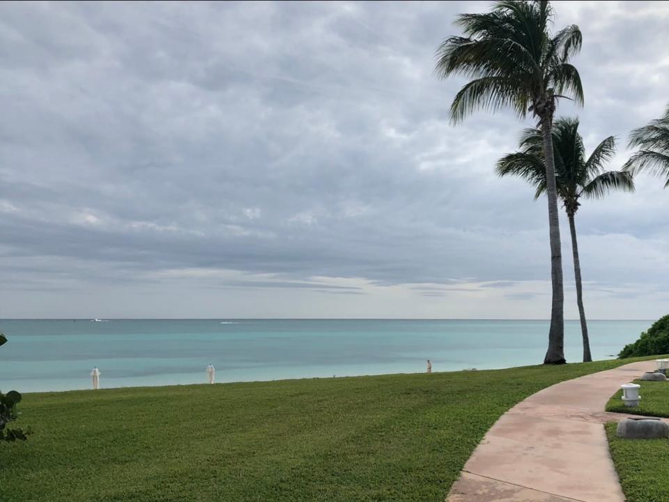 A path, surrounded by greenery and a few palm trees, on a beach. The water is blue and the sky is cloudy.