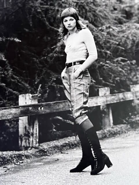 A black-and-white photo of a woman in high black boots, jeans and a T-shirt, standing outside by a low railing.