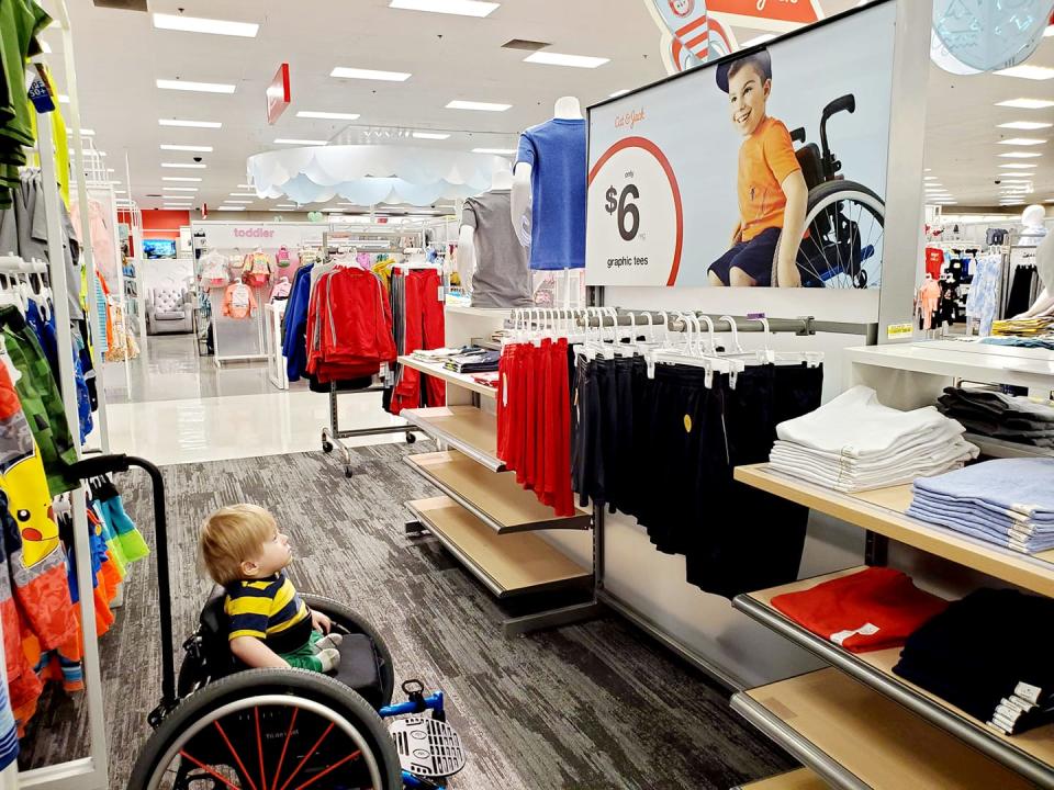 Ollie Garza-Pena, 2, sits in a wheelchair in Target. He's looking at an advertisement featuring a boy also in a wheelchair.