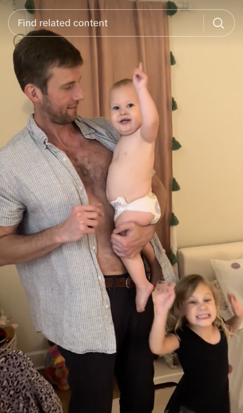 Adult holding a baby in a diaper, with a young child raising their arms in excitement nearby