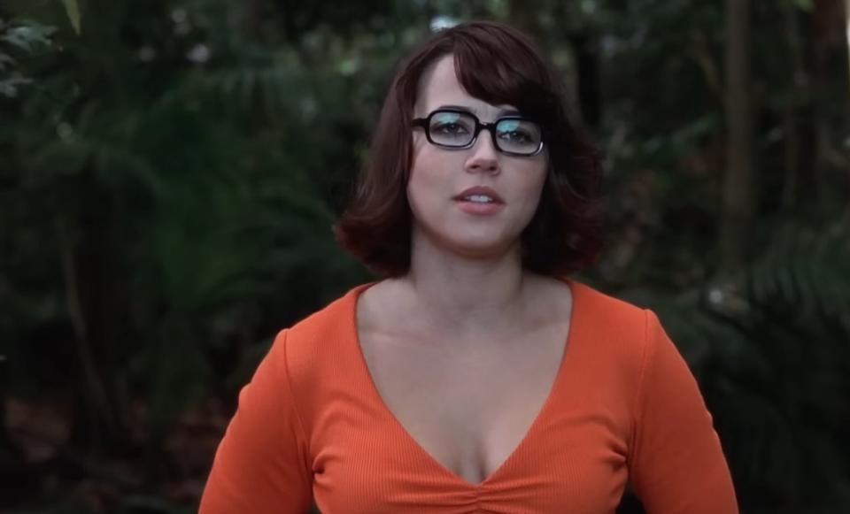 A woman dressed as Velma from Scooby-Doo, wearing glasses and an orange top, stands in a forest