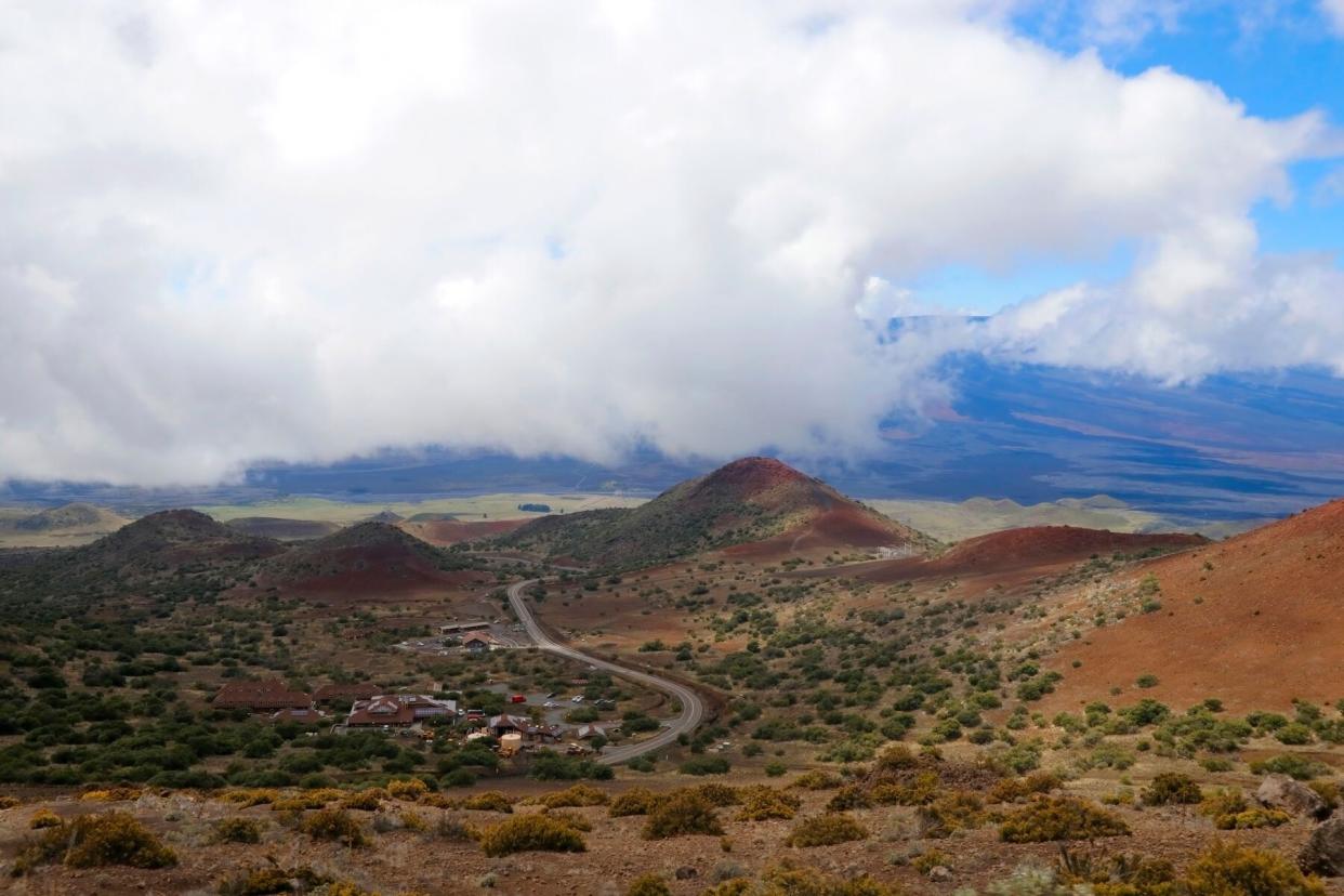 Scenic Mauna Kea landscape with paved road to summit between old craters and view on Mauna Loa in clouds.