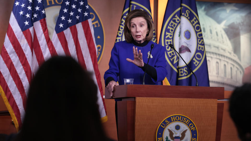 Nancy Pelosi holds out a hand as she stands at a podium with two microphones and a seal that reads: U.S. House of Representatives.