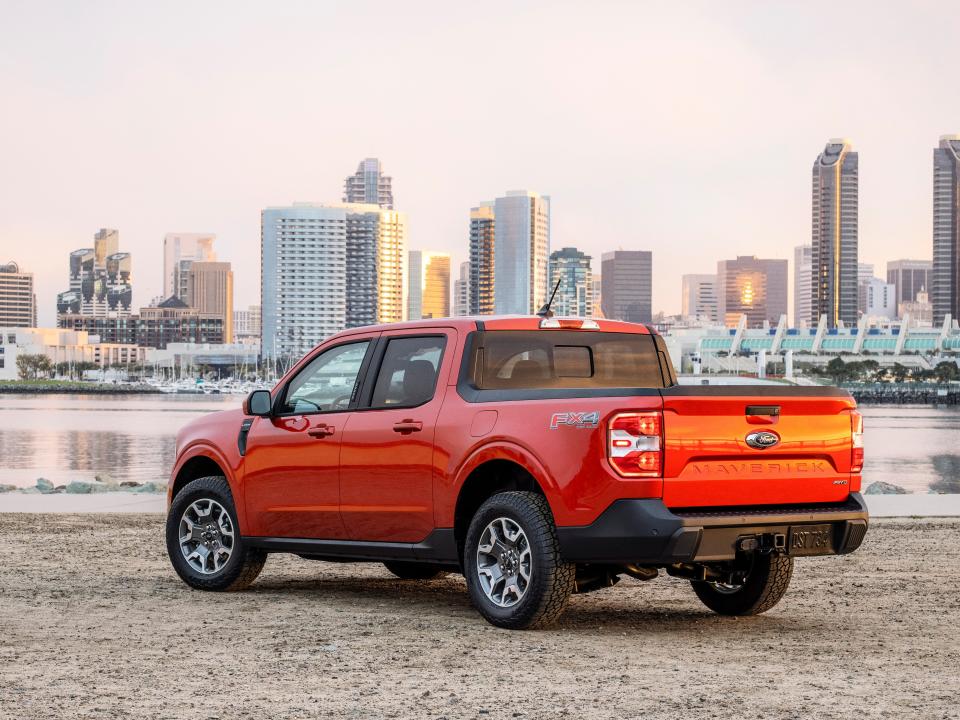 An orange pickup truck is parked in front of a body of water and cityscape.