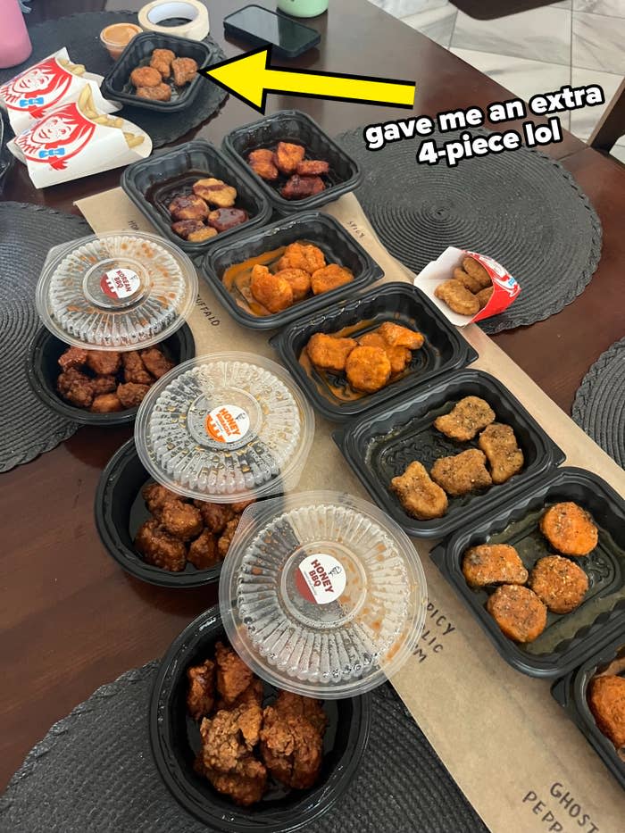 An assortment of chicken wings and nuggets on a wooden table, accompanied by dipping sauces. Black placemats and drink containers are visible