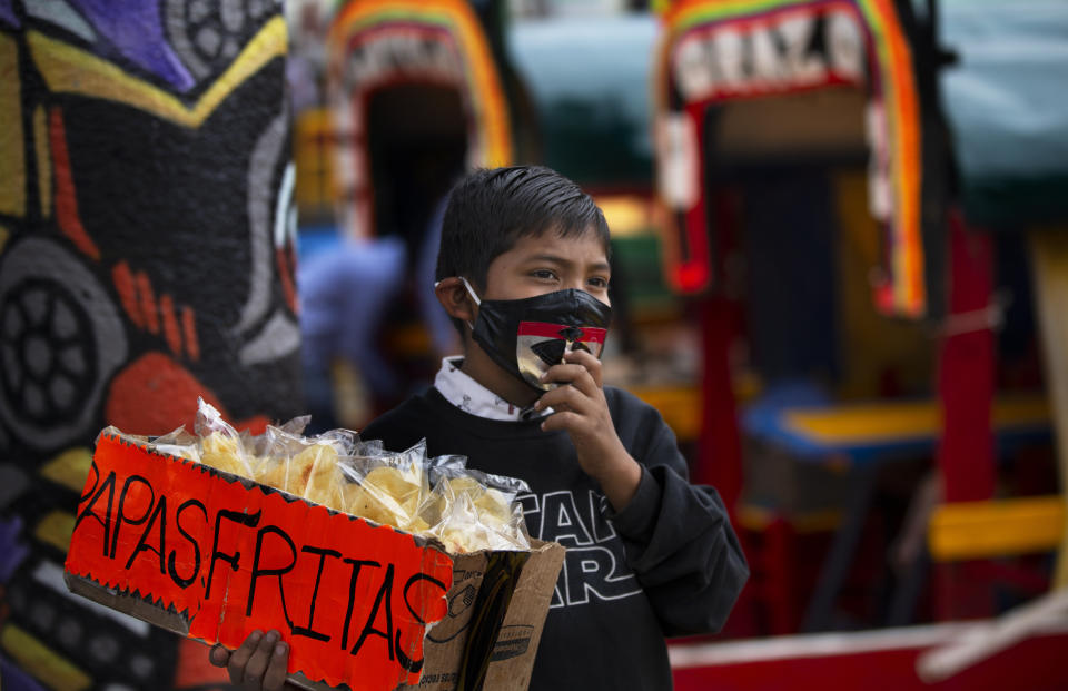 A child, wearing a protective face mask, hawks bags of potato chips near a row of painted wooden boats known as trajineras, popular with tourists that ply the water canals in the Xochimilco district of Mexico City, during a reopening of activities after a six-month pause due to the COVID-19 pandemic. (AP Photo/Marco Ugarte)