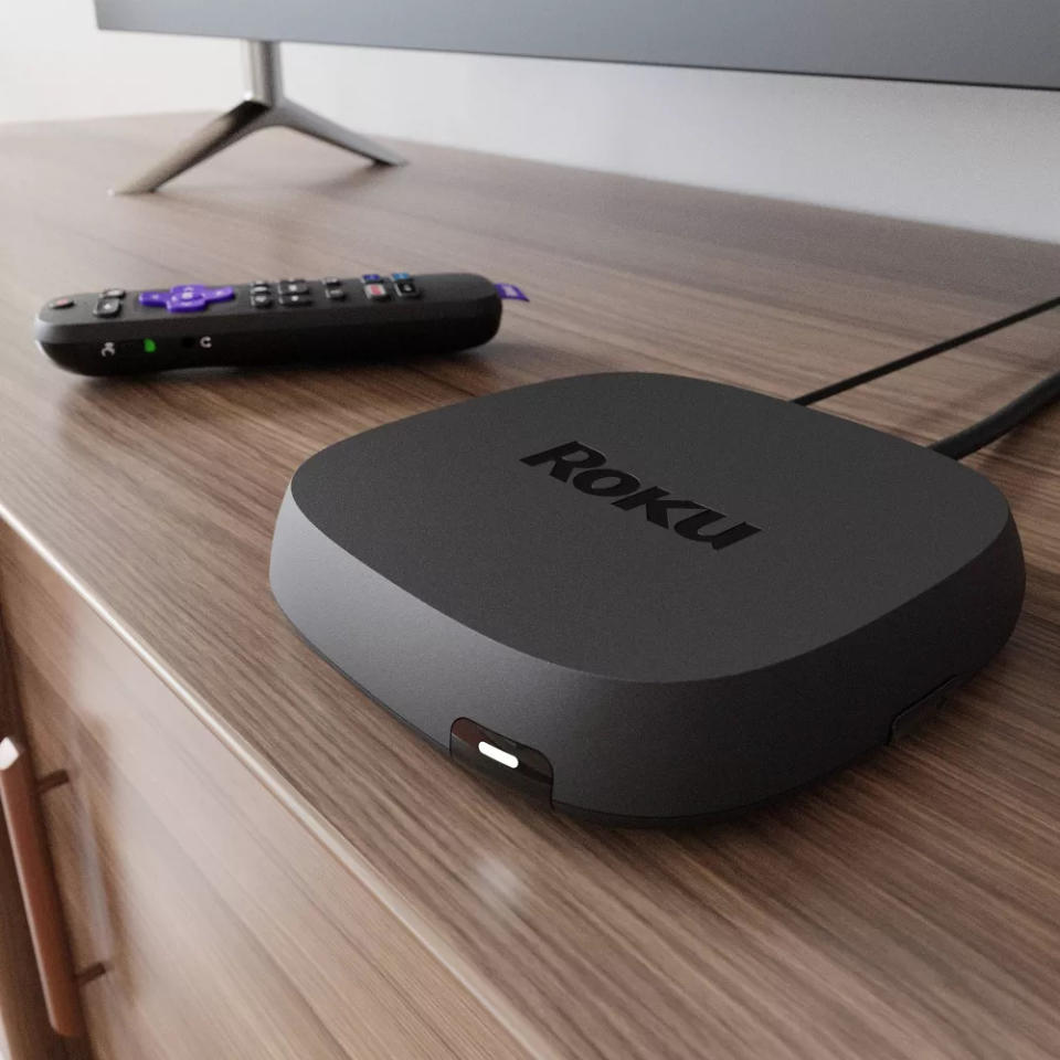 A Roku streaming device sits on a wooden entertainment unit beside a remote control. A TV screen is partially visible in the background