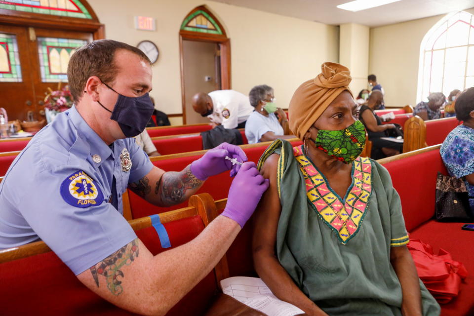 A woman, seated on a church pew, receives a shot in the arm by a paramedic, both wearing face masks