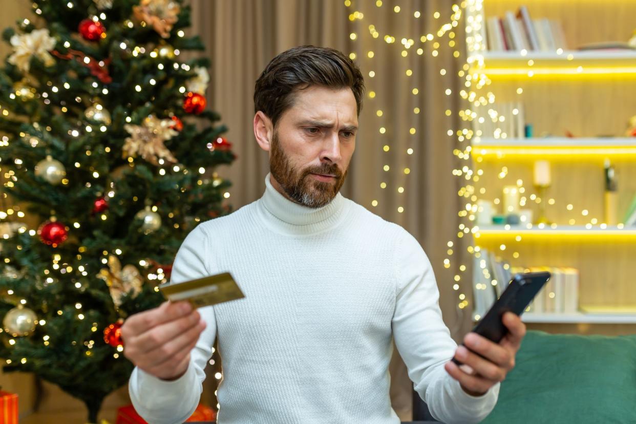 Confused and embarrassed man looks at the phone, holds a credit card in his hands. Sitting on the sofa near the Christmas tree, unable to make festive online purchases, he doesn't have enough money.