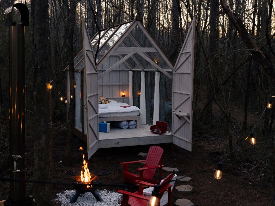 A glass house sits with doors open onto a patio where two red chairs face an outdoor fire.