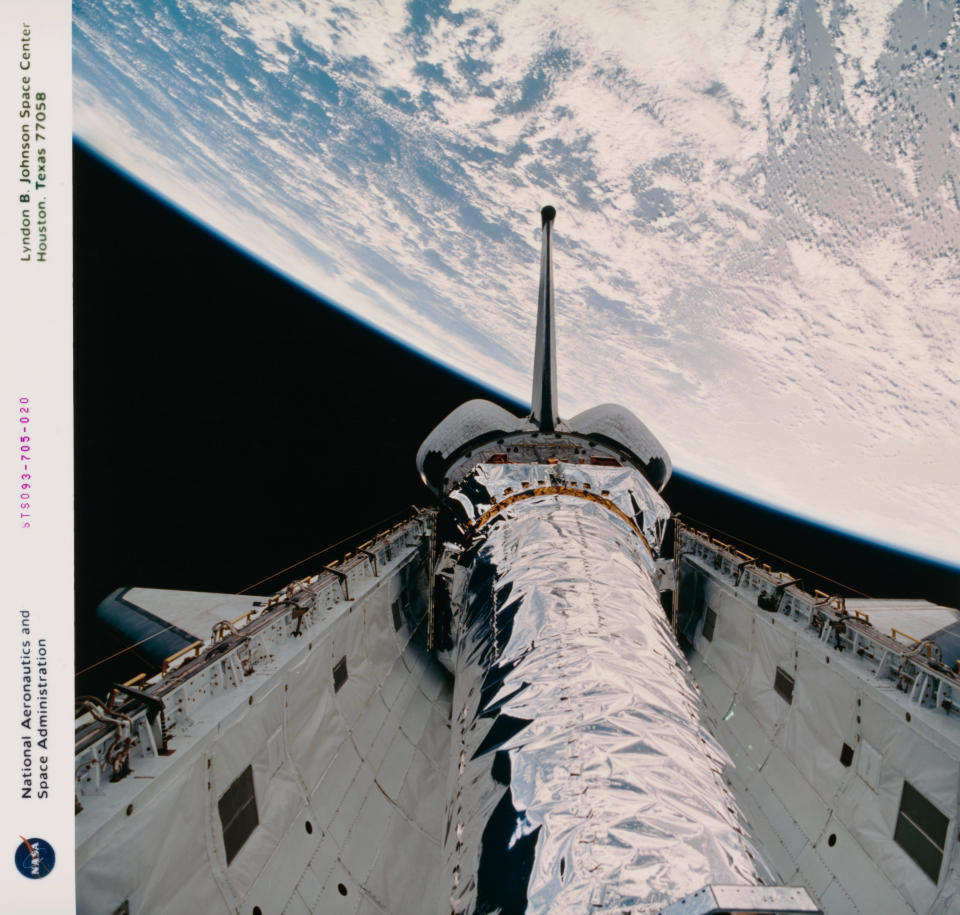 a shiny spaceship rests in the white cargo bay of a space shuttle with a large landmass in the background.