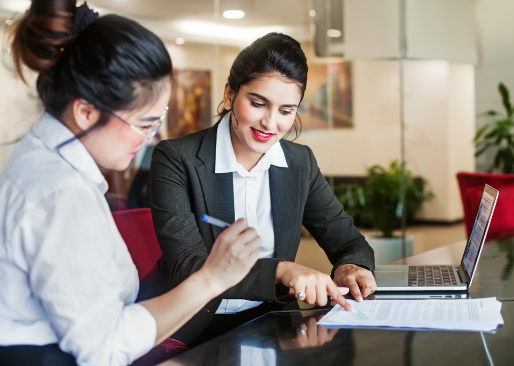 A banker shows a client where to sign while they both sit at a desk in front of a laptop.