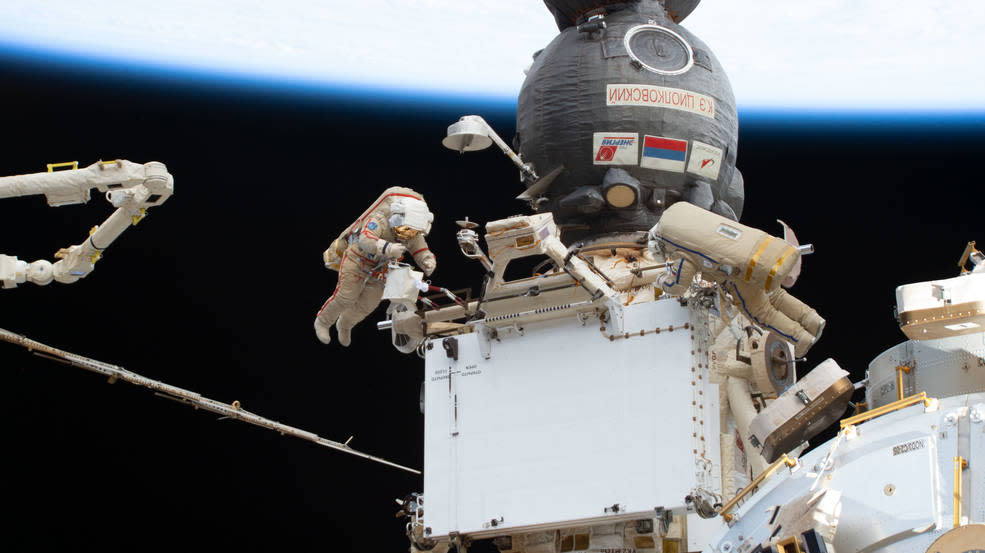  two spacewalkers on either side of an international space station module. each spacewalker wears a spacesuit. at top is a circular module. at left is the edge of the robotic arm. far behind, the curve of the earth 