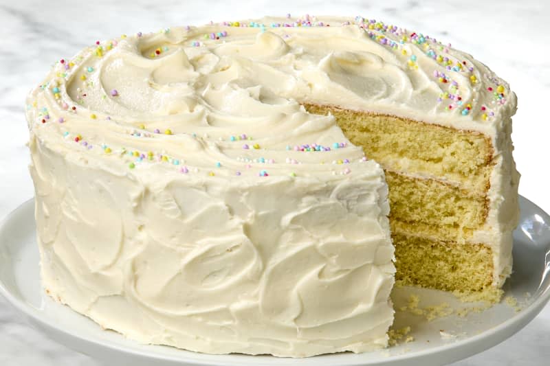 angled shot of a full cake, with a ring of rainbow sprinkles on top, with a slice taken out of it.