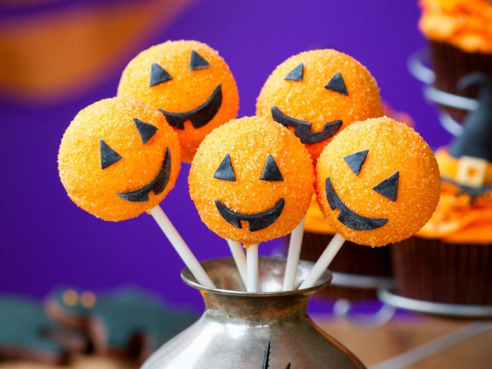 Cake pops decorated like jack-o-lanterns displayed in a silver urn with a plastic spider on it