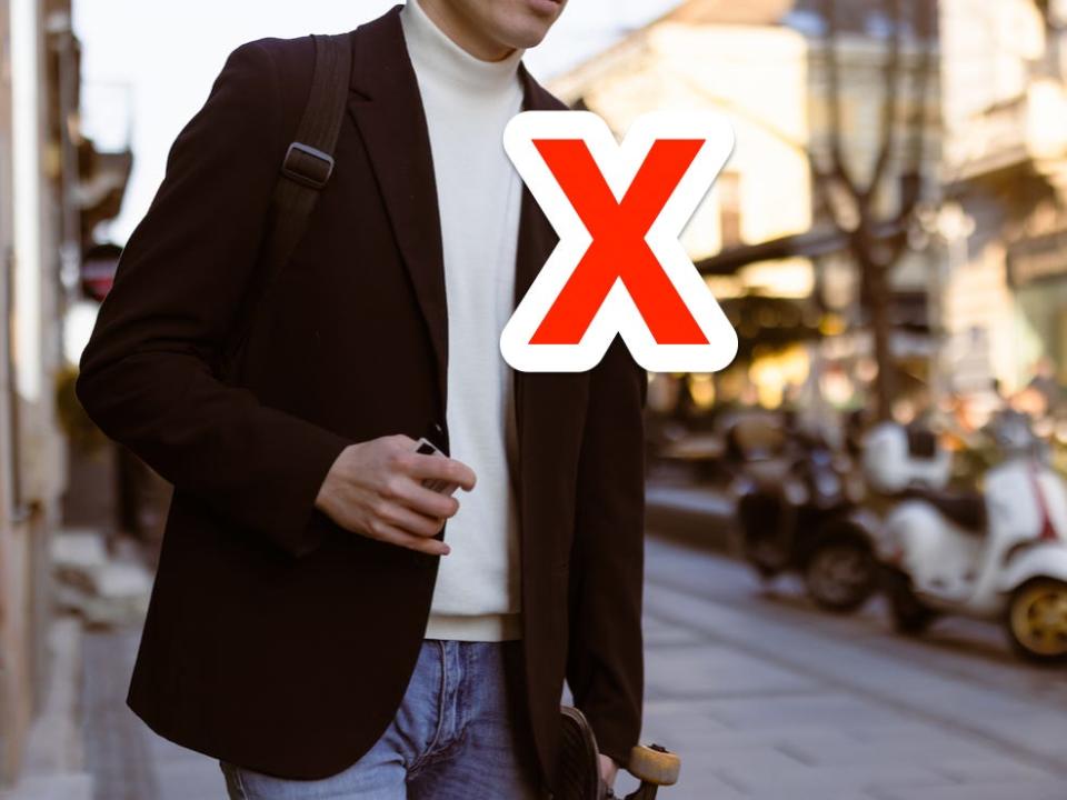 red x on top of person wearing white roll neck mock neck sweater under a brown blazer while carrying a skateboard down the street