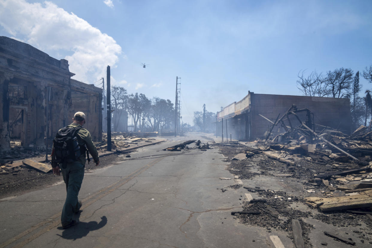 A man walks past smoldering wildfire wreckage on a Lahaina street.