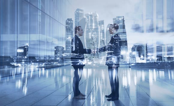 Double exposure of two men shaking hands with office properties behind them.