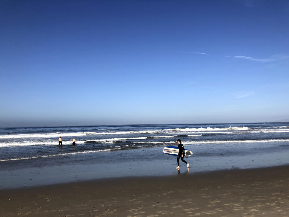 In this Thursday, May 14, 2020, photo surfer Peter Lockwood walks along the beach in Santa Monica, Calif. Masks are required at Los Angeles County beaches, which reopened Wednesday to join counterparts in other states that have allowed a somewhat limited return to famed stretches of sand. (AP Photo/Brian Melley)