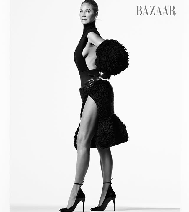 Christy Turlington in a black and white photo for Harper's Bazaar stands in a black dress with a slit with a poofy bottom and elbow pads/poofs