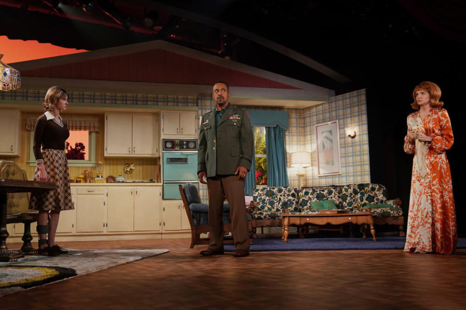 A trio of actors stand on a stage near the set of a 1950's living room