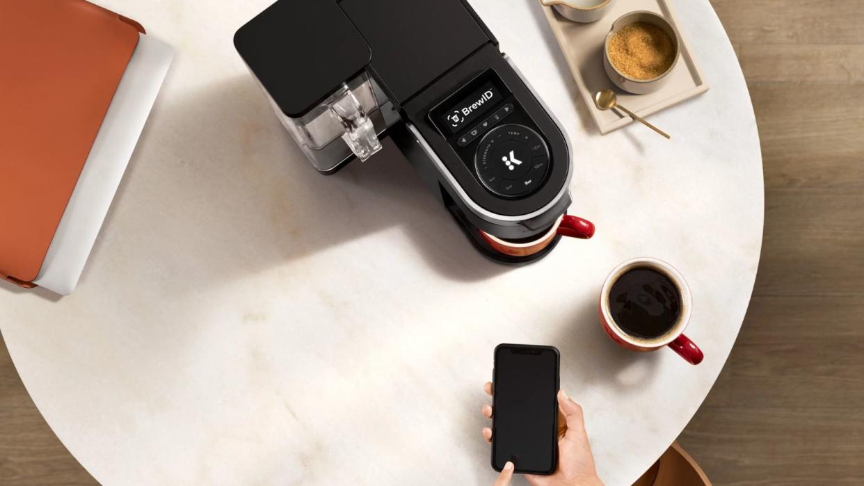  An aerial view of the he Keurig K-Supreme SMART Coffee Maker being controlled by a smartphone next to a cup of coffee. 