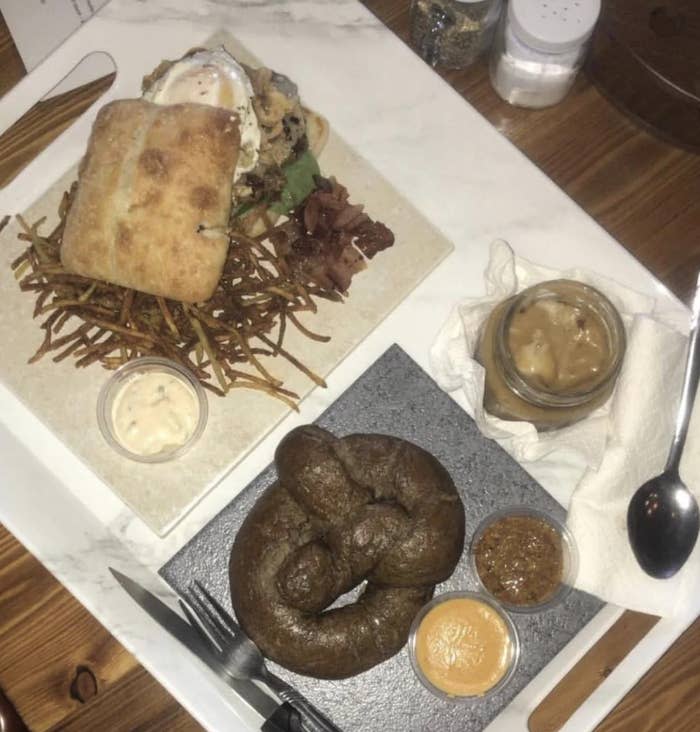 Plated food items include a sandwich on fries, bacon pieces, a glass jar of a brown spread, and a pretzel with dipping sauces. Salt and pepper shakers in the background