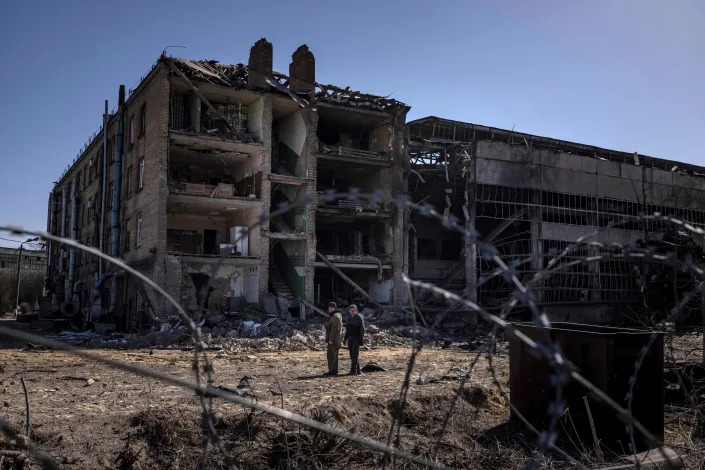 Two people stand beyond a razor-wire fence amid rubble near a partially destroyed building.