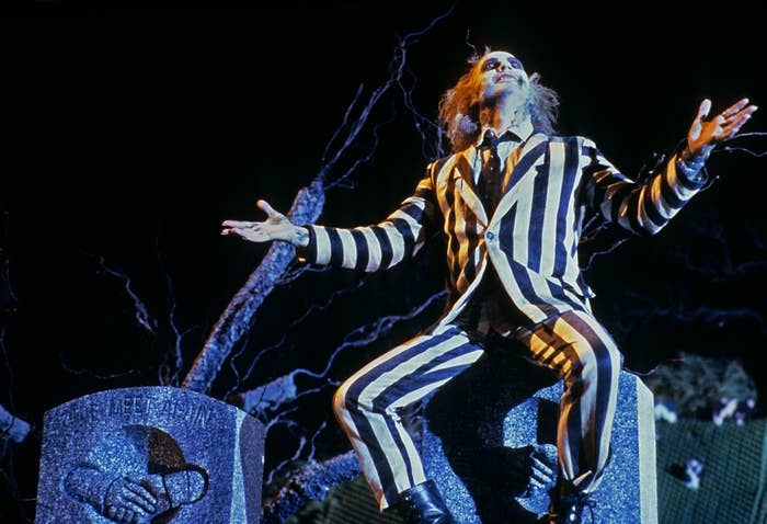Beetlejuice character in striped suit stands with arms outstretched as he sits on a tombstone
