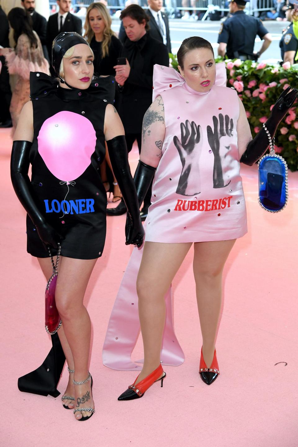 A woman in a black dress with a balloon painted on it and a woman in a pink dress with rubber gloves painted on it stand on a pink carpet.