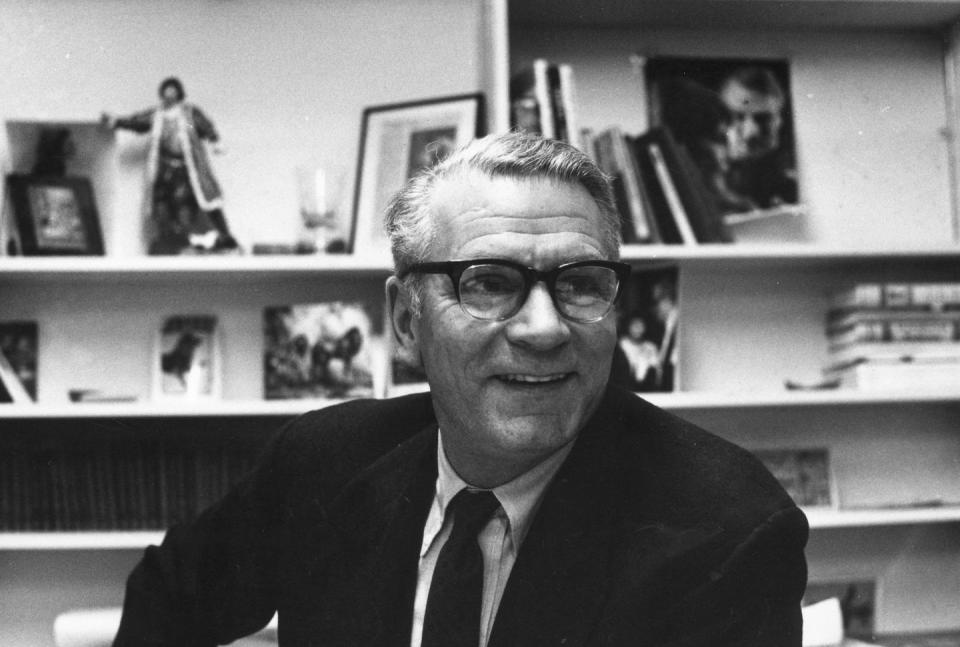 a black and white photo of laurence olivier wearing a suit and tie and glasses, with several bookshelves behind him