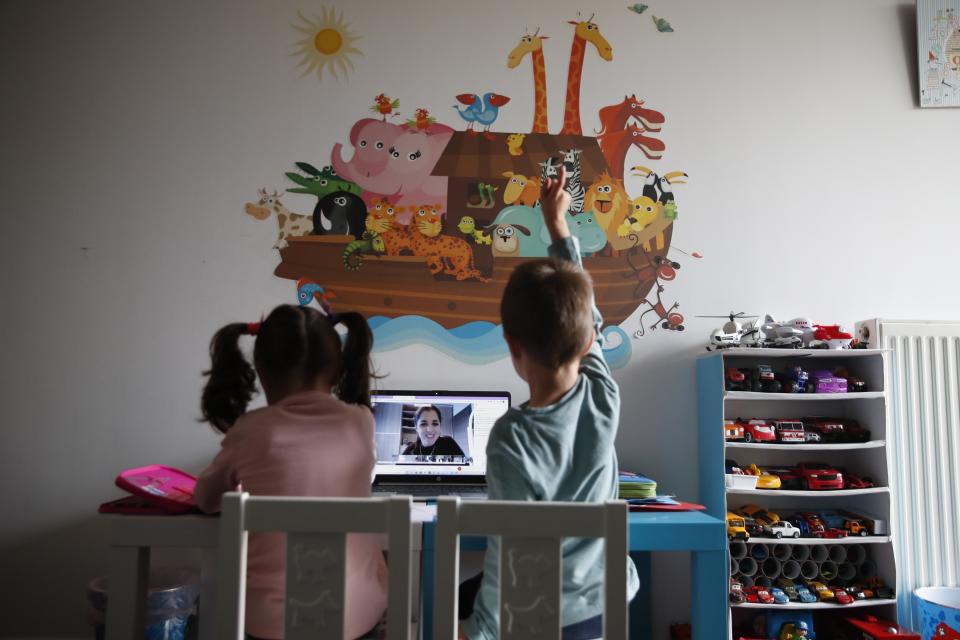 A kindergarten teacher reads a fairy story as Panos raising his hand with his twin sister Marina watch the online lesson in Athens, Thursday, Nov. 19, 2020. Most other European countries have vowed to keep schools open, but the pandemic has hit Greece hard for the first time in recent weeks following a successful lockdown in the spring, overwhelming hospitals in parts of the country. State television is making and broadcasting lessons, while teachers sit in empty classrooms talking to remote students. Despite some problems, they say it keeps children in touch with their schools. (AP Photo/Thanassis Stavrakis)