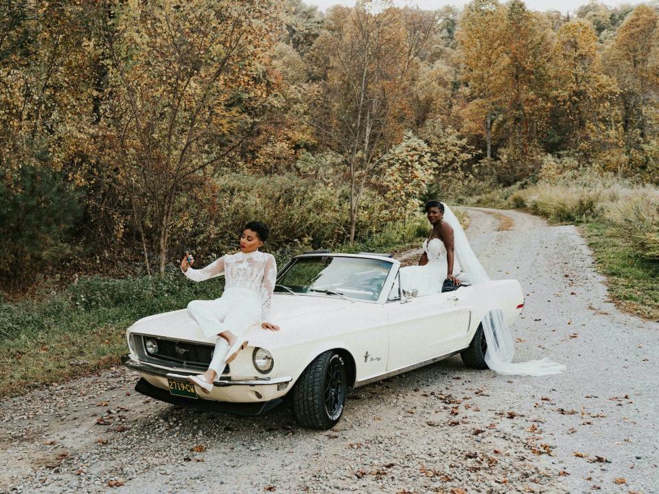 A bride sits on the hood of a cream convertible while smoking a cigar while another bride sits on the back.