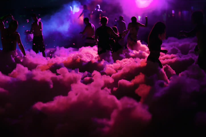 People dance amidst foam and lights at an outdoor event. The image has vibrant lighting but no specific text or recognizable individuals