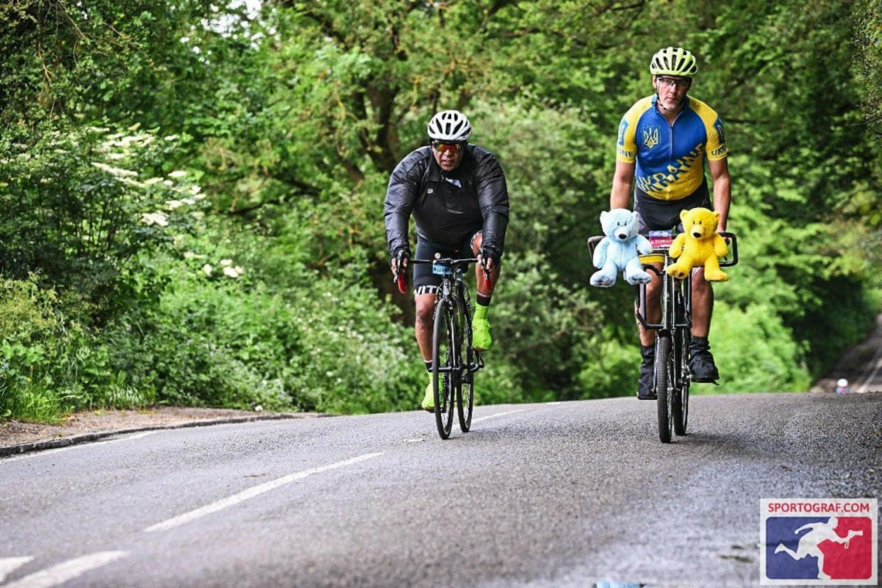 Wide shot of two cyclists where one of the cyclists is wearing a Lycra suit in the colours of the Ukraine flag and teddy bears on his handles