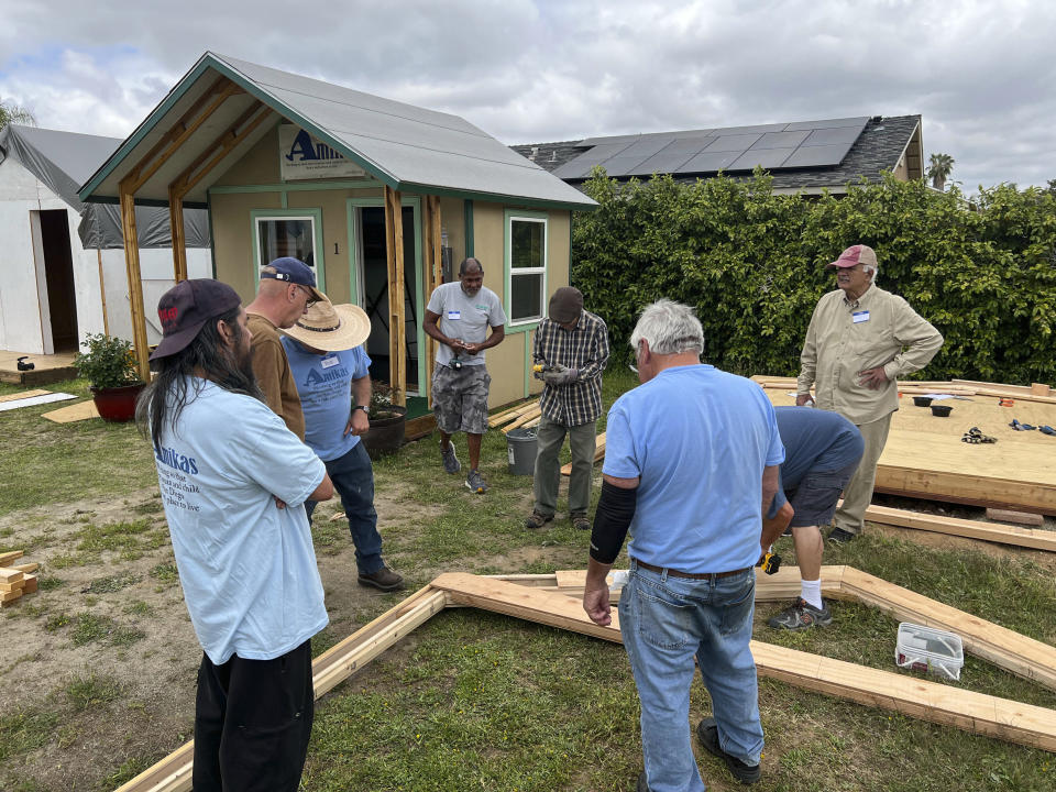 In this photo provided by Amikas, volunteers from all parts of the community gather to assemble the first of seven arches for another cabin at Meridian Baptist Church in El Cajon, Calif., on April 16, 2022. The finished cabin in the background was constructed on site in the fall of 2020 to secure the approval of city officials and the support of the church congregation to host the eventual village of six cabins. The Emergency Sleeping Cabins will serve as bridge housing for homeless single mothers with children. (Lisa Kogan/Amikas via AP)