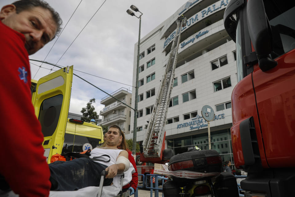 Paramedics carry a man to an ambulance as firefighters are seen on a ladder, some minutes after a fire broke up in the Athenaeum Palace hotel in Athens on Thursday, Dec. 5, 2019. A fire has broken out at a luxury hotel in the Greek capital, with fire crews evacuating the building and using ladders to rescue several people.(AP Photo/Petros Giannakouris)