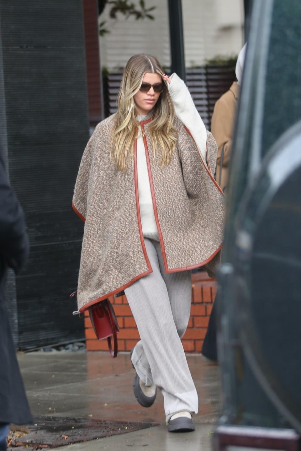 Sofia Richie Grainge wearing a leather-trimmed neutral-toned cape, ribbed knit pants, and rubber boots out on a rainy day