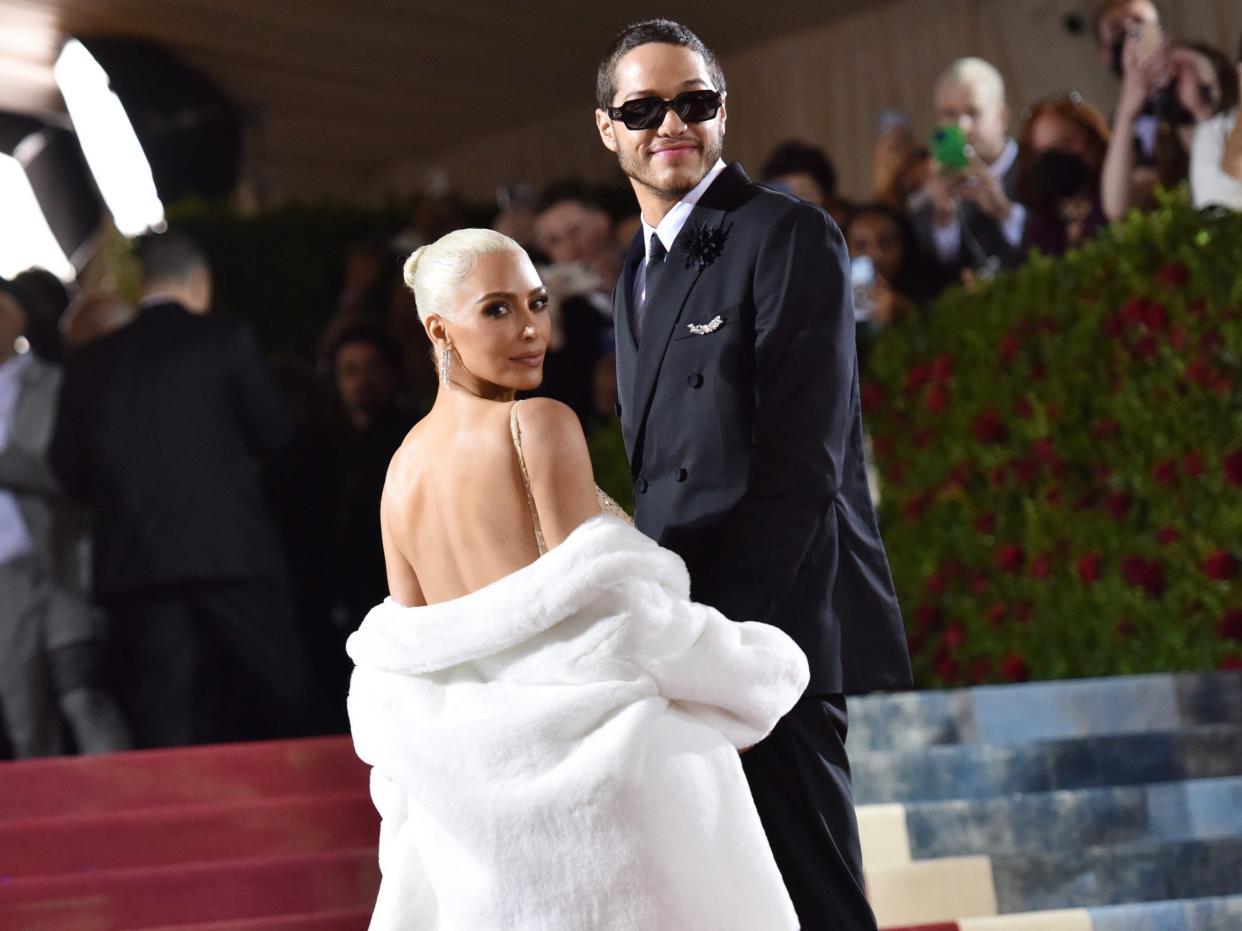 kim kardashian and pete davidson on the steps at the met gala, looking towards the side. kardashian is looking over her shoulder, a white fur coat hanging around her elbows, and davidson is wearing sunglasses