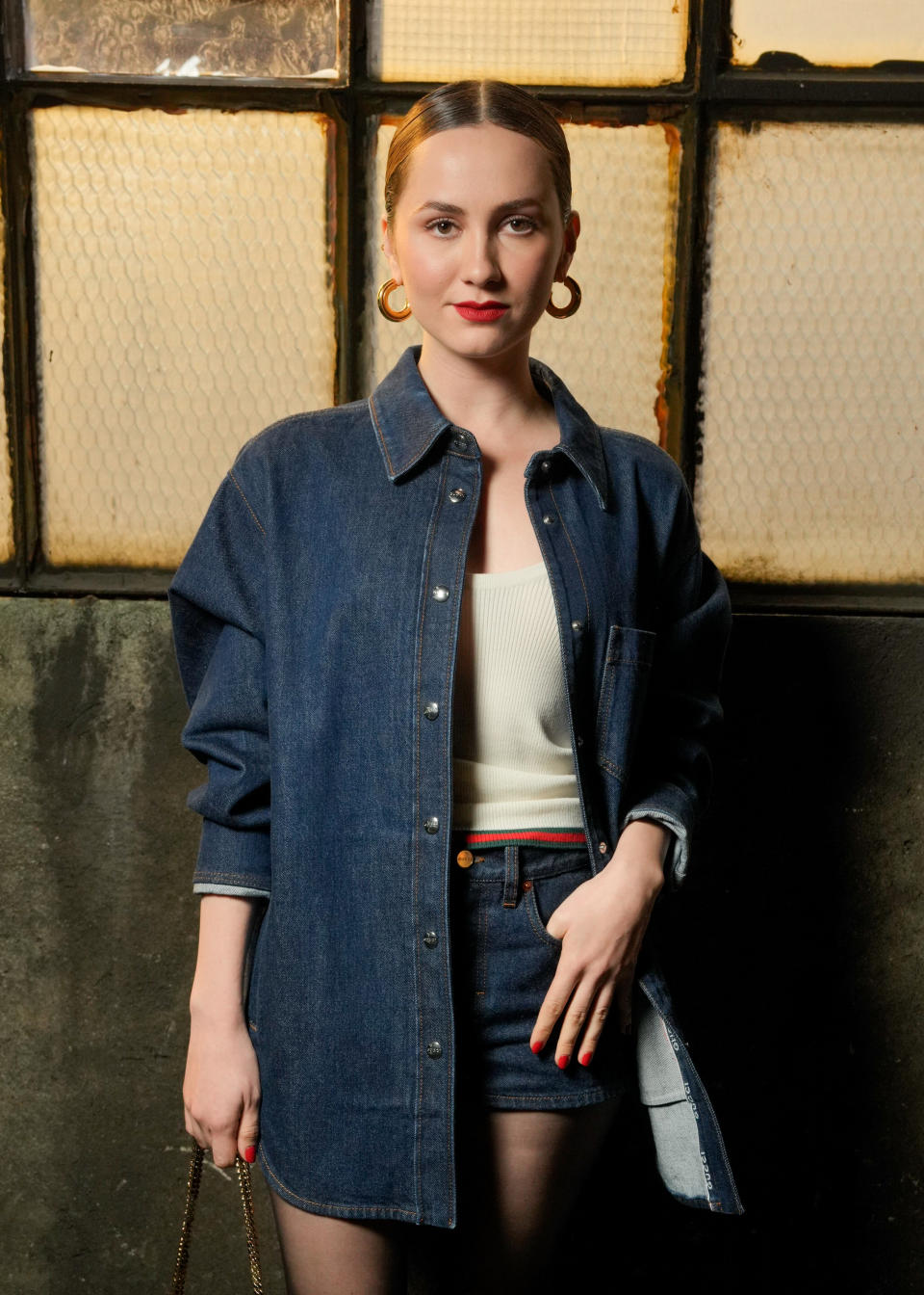 A woman in a stylish, oversized denim button-up shirt and denim shorts, accessorized with large gold hoop earrings, stands confidently in front of a textured window