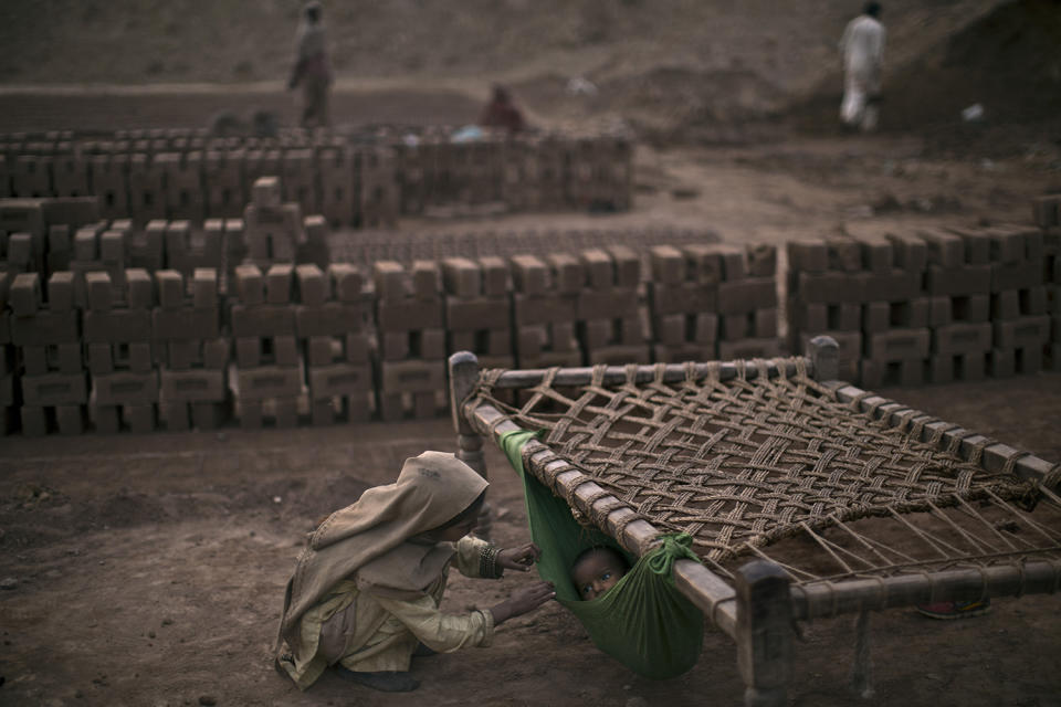 <p>Pakistani girl, Kiran Riasat, 8, who works with her mother and father, seen in the background, in a brick factory, checks on her brother, Rizwan, 1.5, laying in a hammock attached on a bed, at the site of work, in the outskirts of Islamabad, Pakistan, Feb. 18, 2014. (Photo: Muhammed Muheisen/AP) </p>