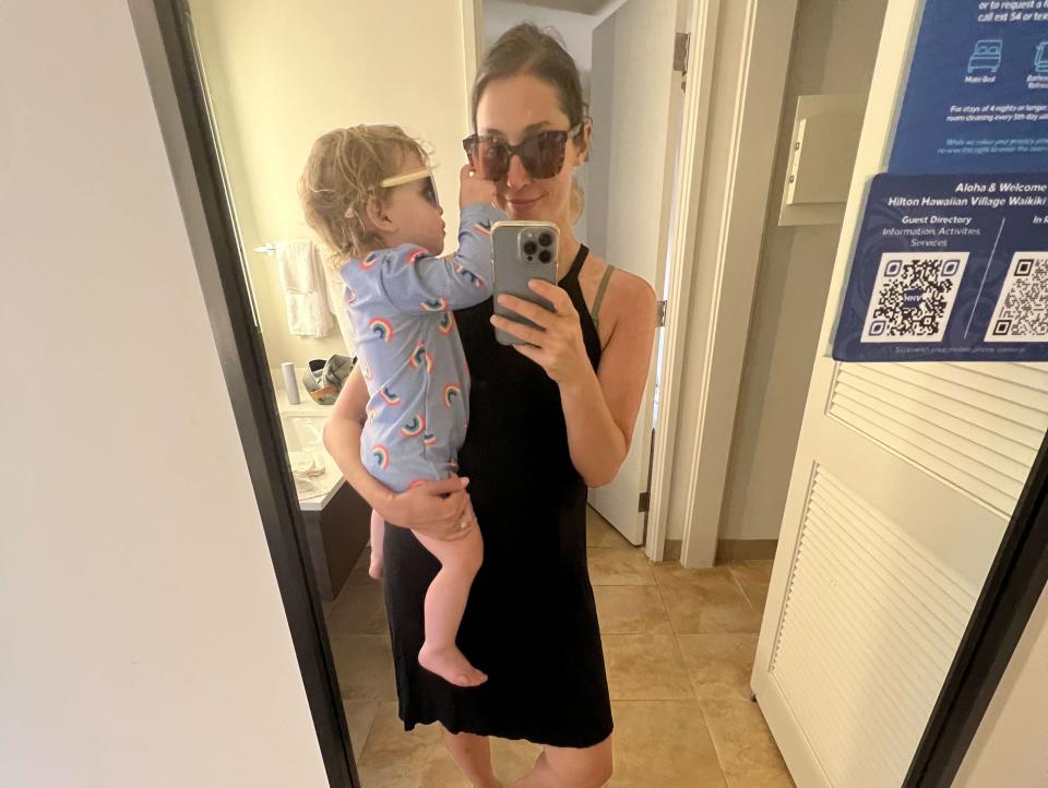 A woman taking a mirror selfie holding her baby who is pointing to her face and both are wearing sunglasses.