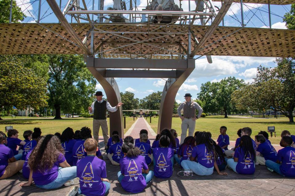 Elementary students from Bear Exploration Center For Mathematics, Science and Technology School perch in the shade of the Wright Flyer replica as they learn about the history of aviation from “Orville” and “Wilbur Wright,” May 13, 2022.