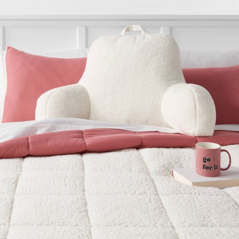 Fluffy backrest pillow on bed with pink pillows, comforter, and 'go for it' mug. Ideal for cozy reading nook