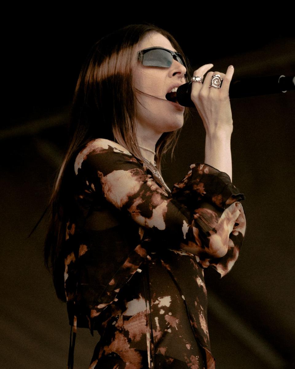 a close up of caroline polachek singing in a brown burnout top, wraparound sunglasses, and silver rings