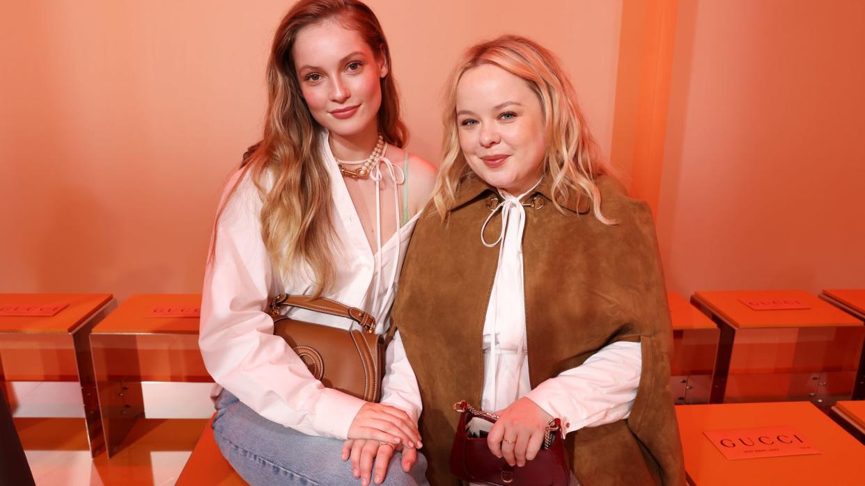 Hannah Dodd and Nicola Coughlan sitting together at the Gucci Women's Spring Summer 2025 Fashion Show. Both look chic—Hannah in her casual jeans and white shirt, and Nicola in her brown cape and white dress. They both sport pointed heels, Hannah's in burgundy and Nicola's in green. The two are smiling, appearing relaxed and engaged.