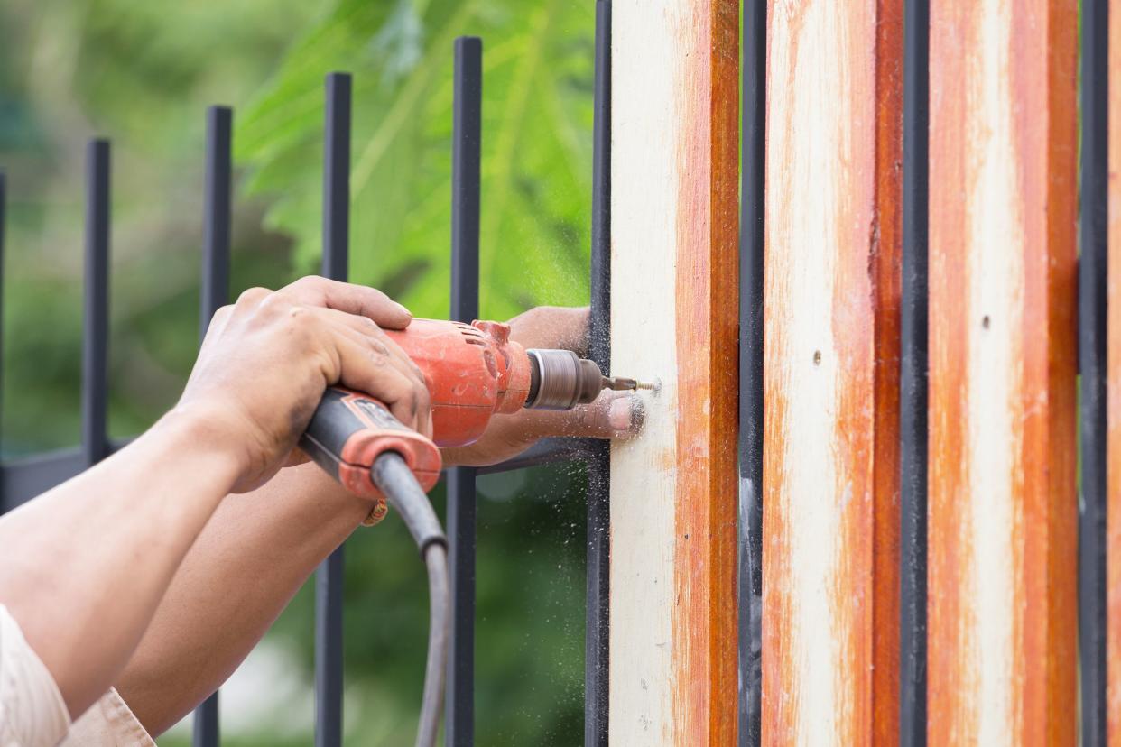 Male carpenter hands using electric drill to drill pieces of wood on fence with blurred background of tree foliage