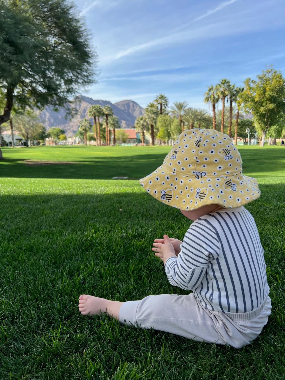 A child wearing a yellow hat sits on the grass with the mountains in the distance.