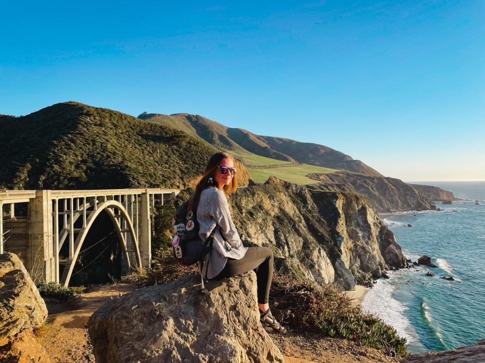 Emily sits on a rock in front of Bixby Bridge along the coast in Big Sur. The water is blue and mountains are green.